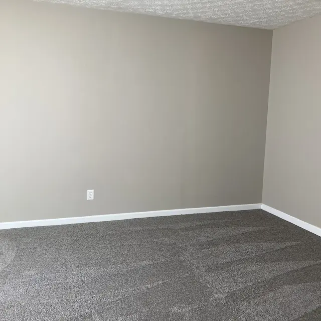 An empty room with light gray walls and plush gray carpet. The room has no furniture and a neutral aesthetic, featuring a ceiling with a textured finish.
