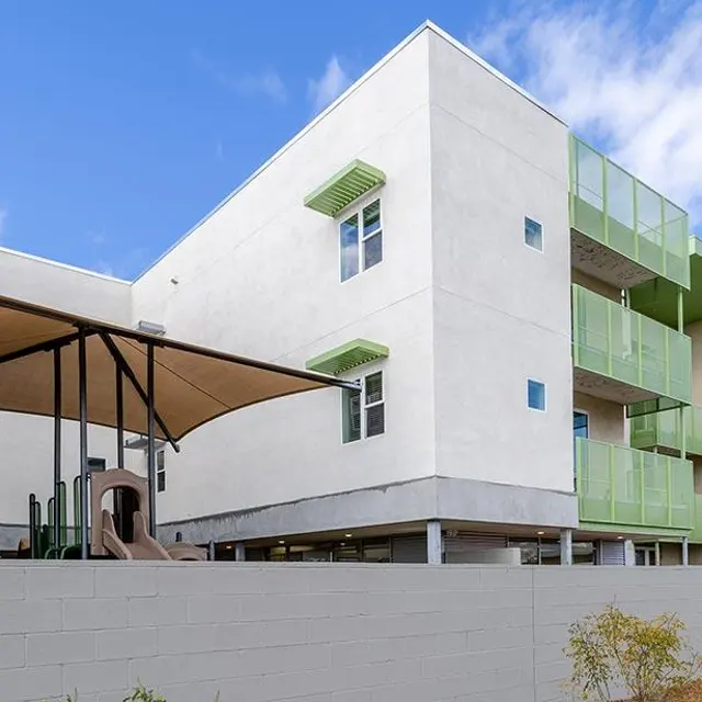 A modern apartment building featuring a playground area with a shade structure. The building has multiple stories and green accents on the balconies.