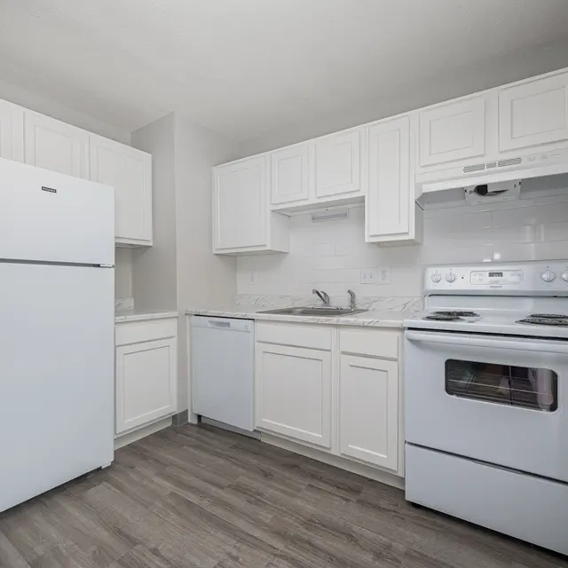 A modern kitchen featuring white cabinetry, a white refrigerator, a white dishwasher, and a white stove. The floor is made of gray laminate wood, creating a bright and spacious environment.