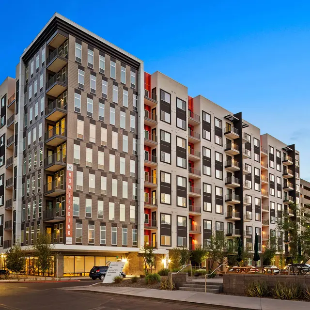 A modern multi-story apartment building at dusk with a blue sky. The building features a contemporary design with an array of balconies and large windows.