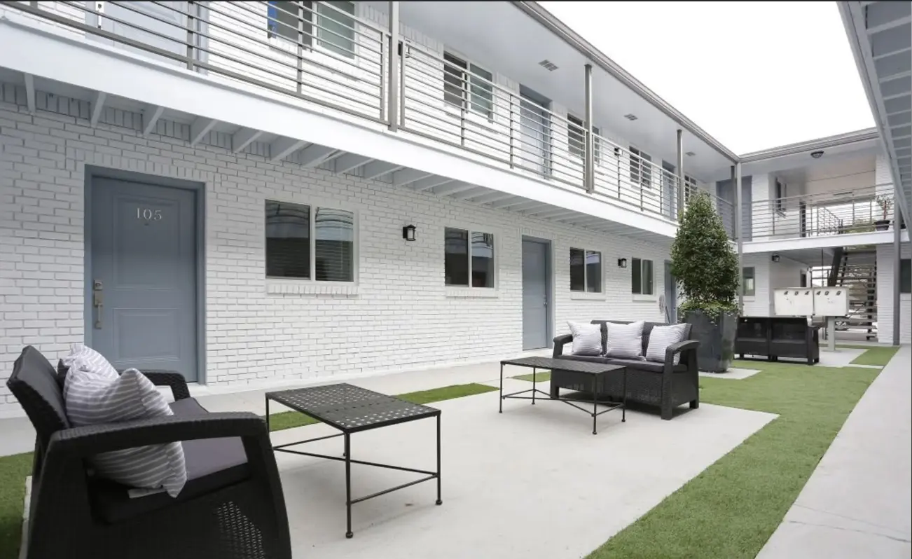 A modern patio area of an apartment complex featuring lounge chairs, a coffee table, and green grass under a cloudy sky.