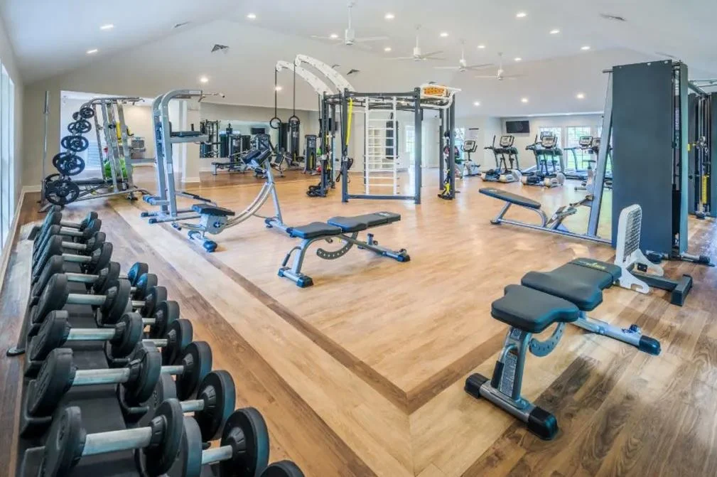 A spacious gym interior featuring various exercise equipment including dumbbells, weight machines, and cardio machines on wooden flooring.