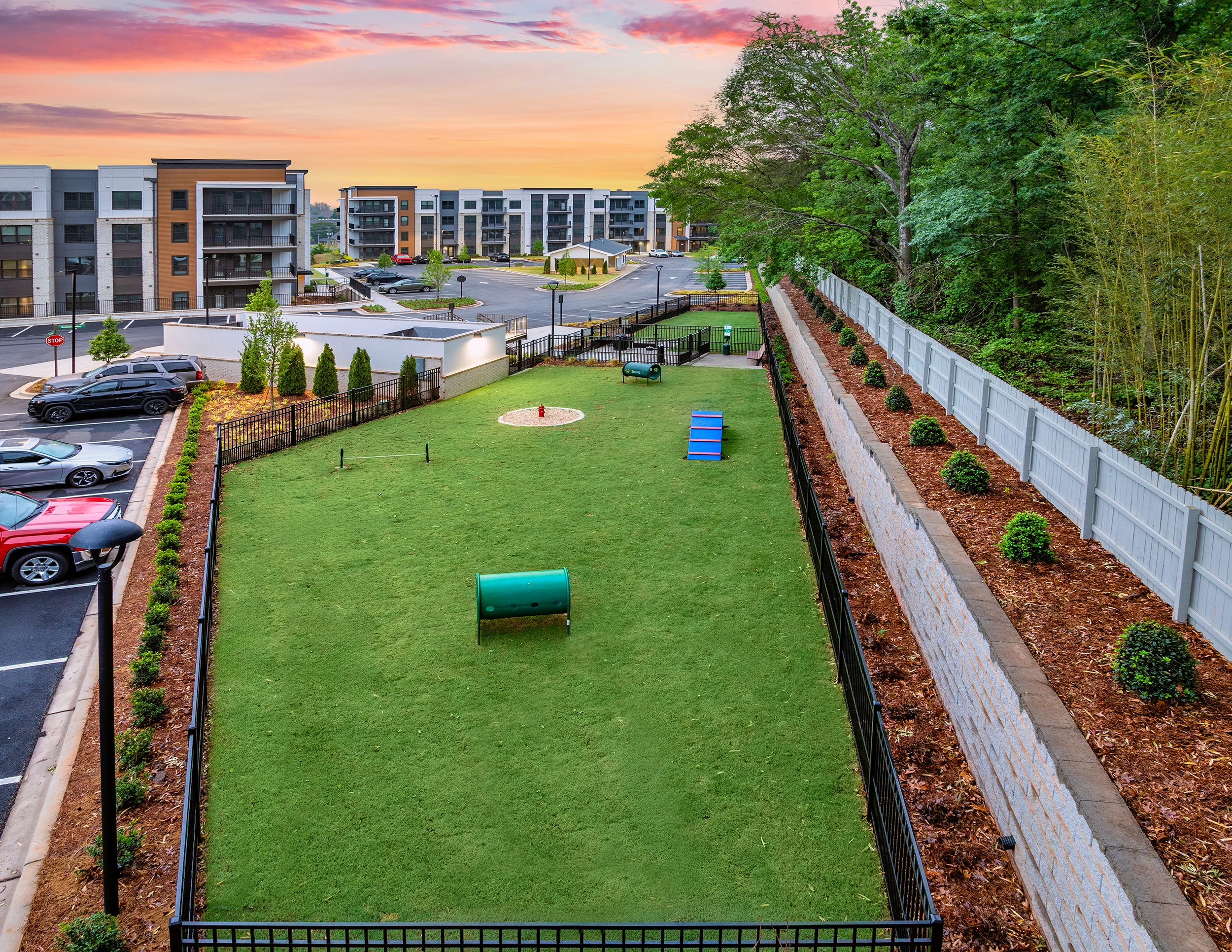 A landscaped dog park with a grassy area and play equipment, surrounded by a black fence and residential buildings in the background under a colorful sunset.