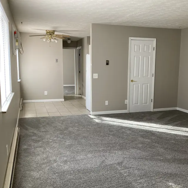 A spacious interior living room with gray carpet and light-colored walls. A ceiling fan hangs from the ceiling, and there are windows allowing natural light to flow in. A door is visible on the right, and there is a tiled area on the left, leading to another room.