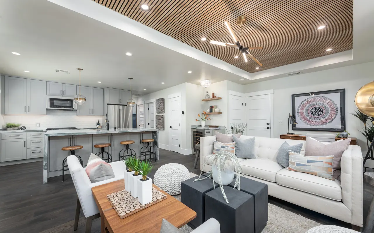 A modern and stylish living room with a light color palette and wooden ceiling. It features a white sofa with decorative pillows, a coffee table with decorative items, bar stools at the kitchen island, and a contemporary kitchen in the background.