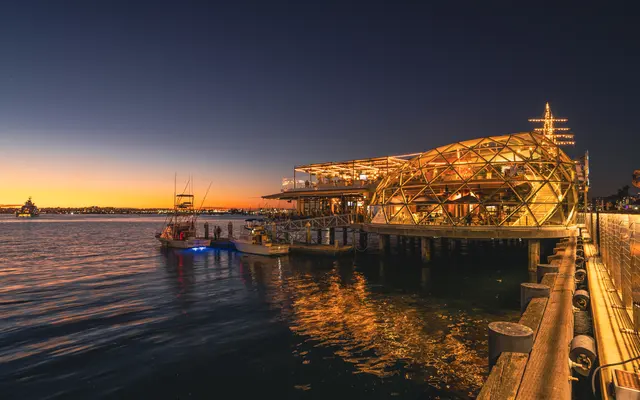 A scenic view of a waterfront structure illuminated at sunset, with boats docked nearby and a calm water surface reflecting the golden hues of the evening sky.