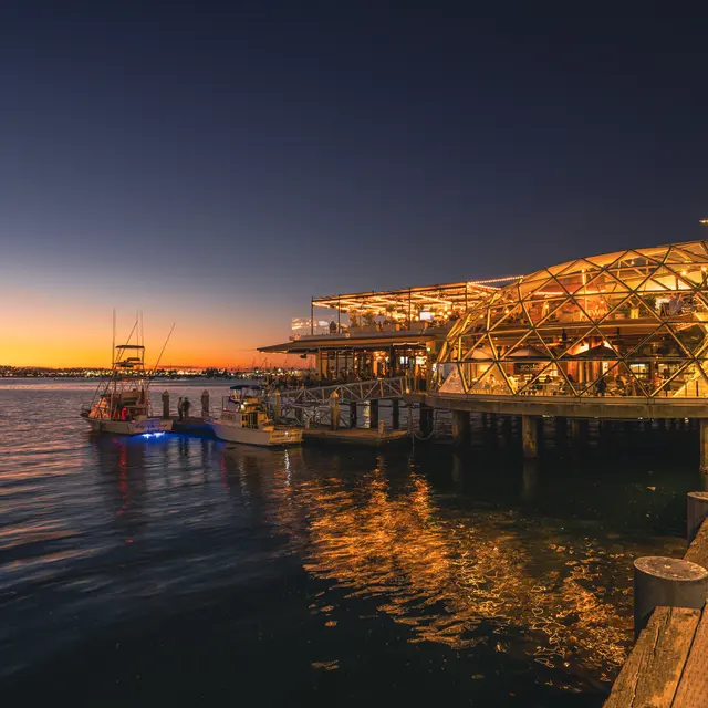 A scenic view of a waterfront structure illuminated at sunset, with boats docked nearby and a calm water surface reflecting the golden hues of the evening sky.