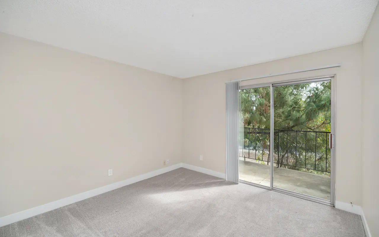 An empty living room with beige walls and carpet, featuring a sliding glass door leading to a small balcony.
