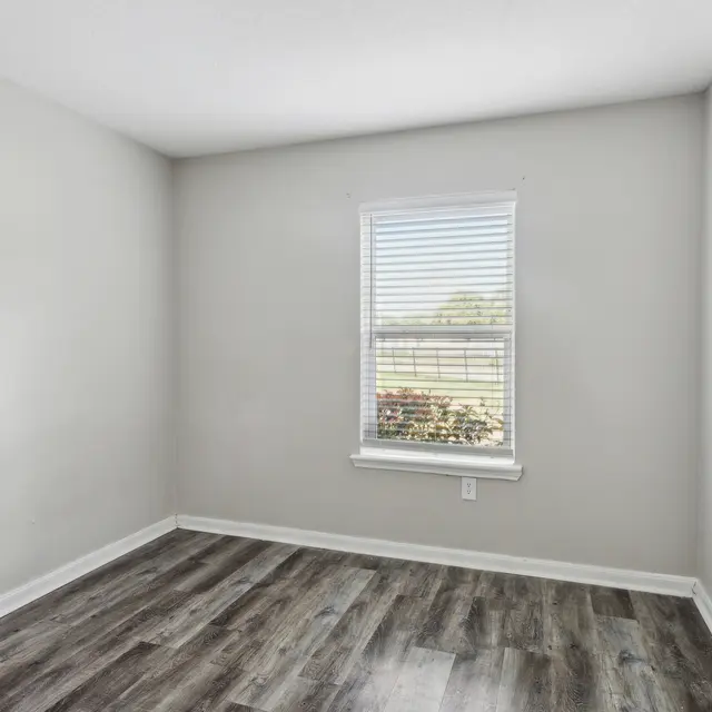 Empty Room Interior An empty room with light gray walls and wooden flooring. A single window with white blinds lets in natural light.