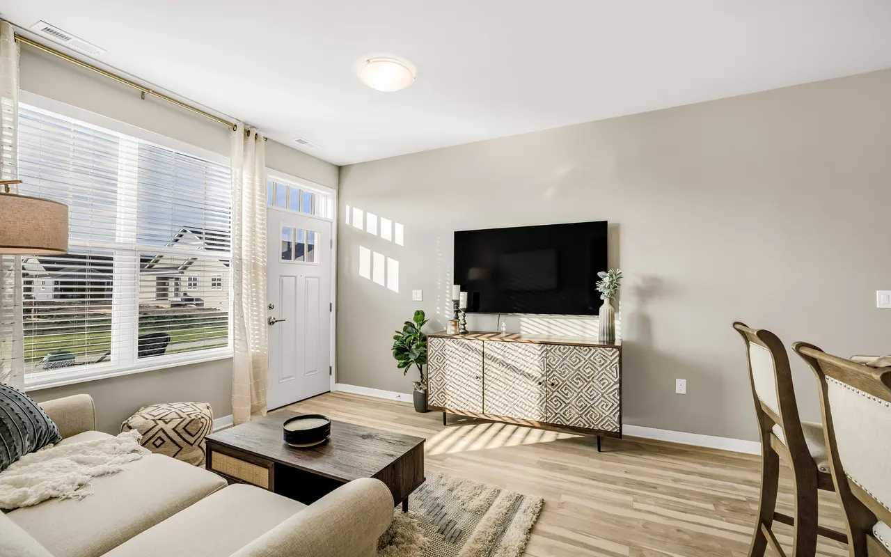 A contemporary living room featuring a light beige sofa, a wooden coffee table, and a TV mounted on the wall. Sunlight streams through large windows adorned with white curtains, illuminating a patterned rug and a decorative cabinet. Wooden flooring complements the modern decor.
