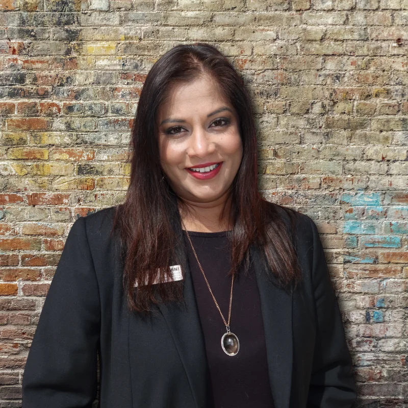 Portrait of a Woman in a Black Jacket A woman smiling, wearing a black jacket, with long dark hair, standing against a textured brick wall.