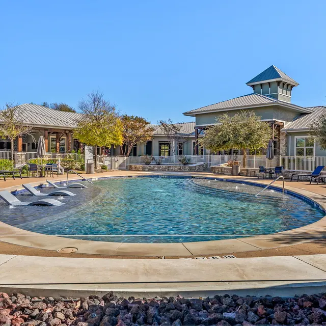 View of a swimming pool area with lounge chairs and a clubhouse in the background on a sunny day.