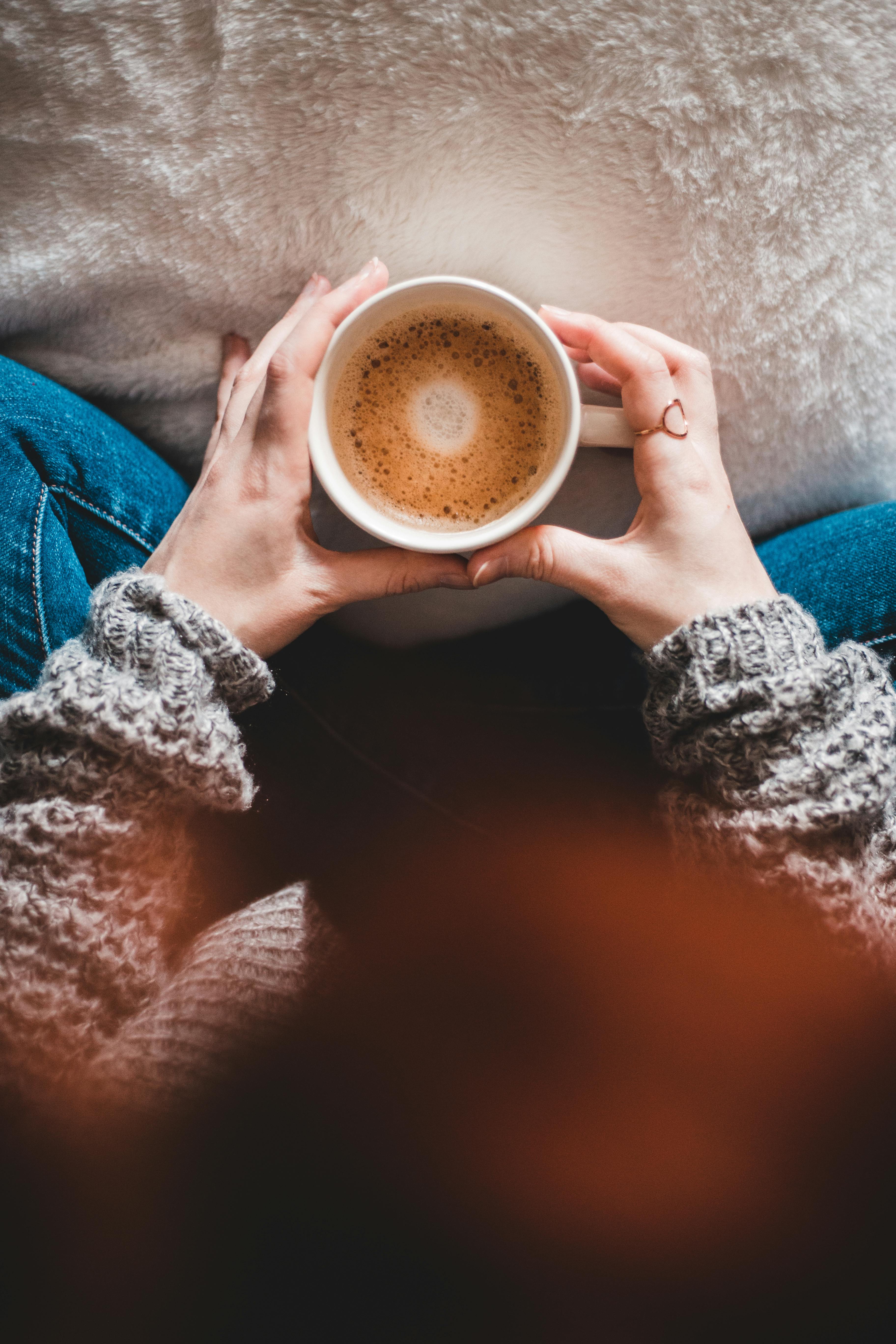 Cozy Coffee Moment A top-down view of a person holding a white coffee cup with frothy coffee, wearing a cozy grey sweater, seated on a light-colored surface.