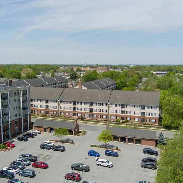 Aerial view of a parking lot with various cars parked, surrounded by buildings including a multi-story apartment complex and low-rise structures. A water tower is visible in the background. The landscape consists of green trees and open areas.