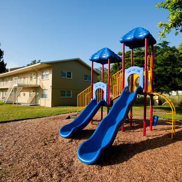 A colorful playground featuring two blue slides, set in a grassy area with wood chips, alongside a multi-story apartment building under a clear blue sky.