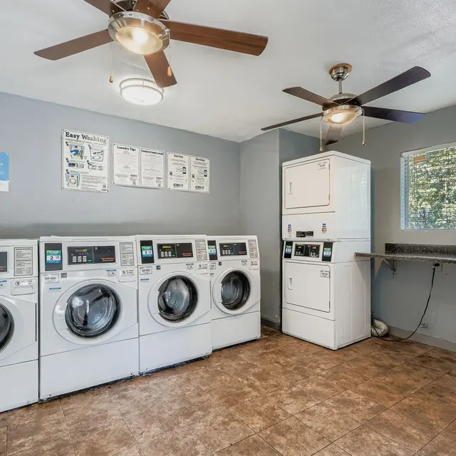 Interior view of a laundry room featuring four front-loading washing machines, a dryer, and a folding table, with two ceiling fans and a window letting in natural light.