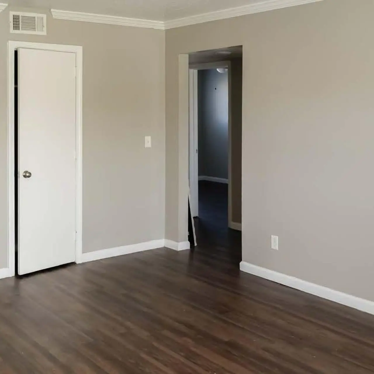 A spacious living room area with light brown hardwood floors and light grey walls. There is a closed white door on the left side, and an open doorway leading to another room in the background. A light source coming from the window on the left illuminates the space.