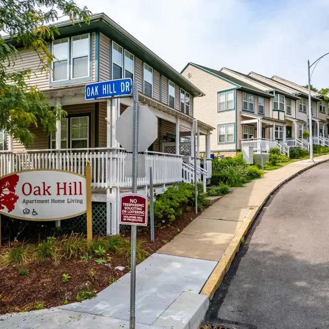 Oak Hill Apartment Complex A residential building complex with a sign showing 'Oak Hill Apartment Home Living'. The houses are arranged on a sloped street, surrounded by greenery and trees.