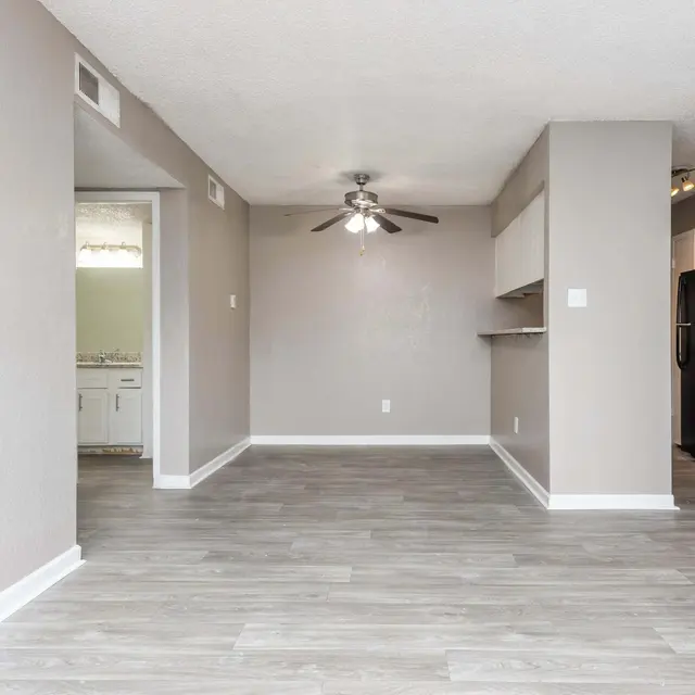 A modern living area with light gray walls and hardwood flooring, featuring a ceiling fan and open space connecting to a kitchen area with appliances.