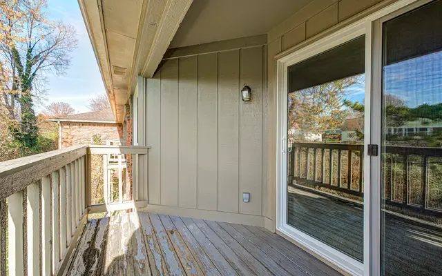 A wooden balcony with a weathered floor and a modern sliding glass door. The walls are painted light beige, and the shot captures the natural surroundings outside the balcony, including trees in the distance.