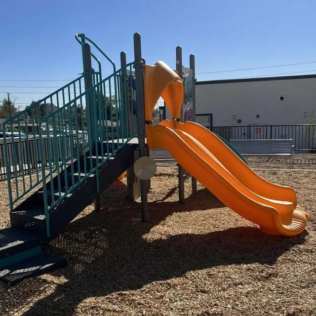 An outdoor playground structure featuring two orange slides connected to a set of blue stairs. The area is covered in wood chips and there are clear blue skies overhead.