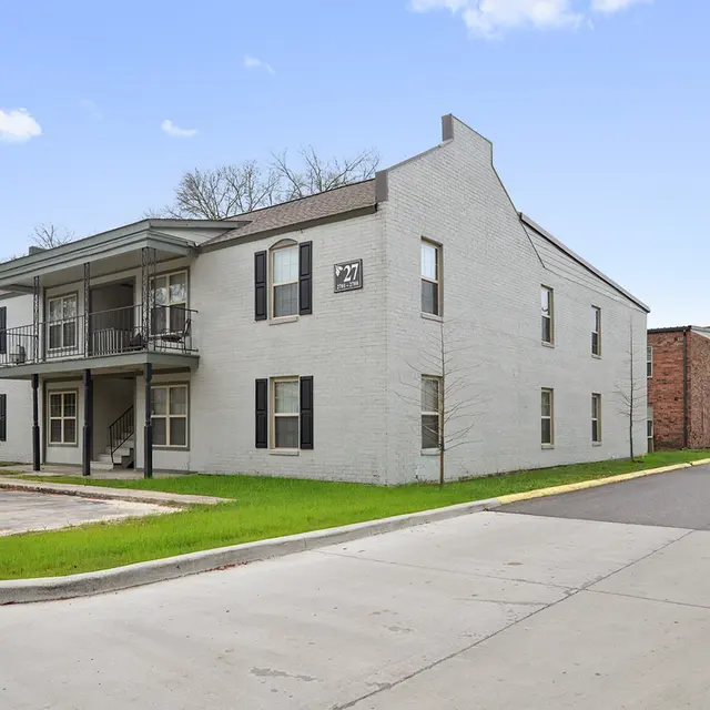 Exterior view of a two-story apartment building with a light gray facade and small balcony, surrounded by green grass and a paved road.
