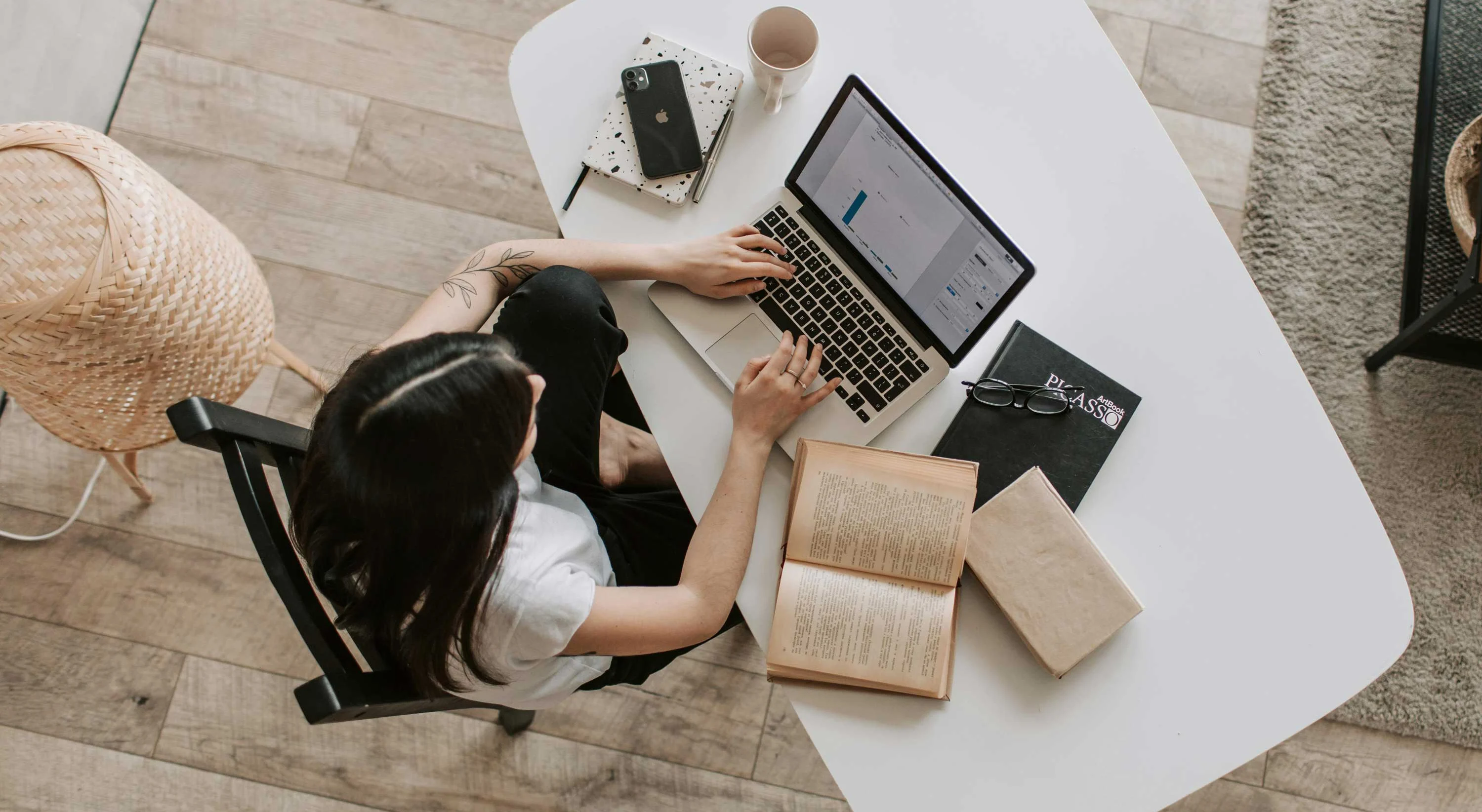 Cozy Workspace with Laptop and Books A woman sitting at a white desk with a laptop, books, and a notebook, engaged in work. The desk is surrounded by a cozy environment with wooden flooring.