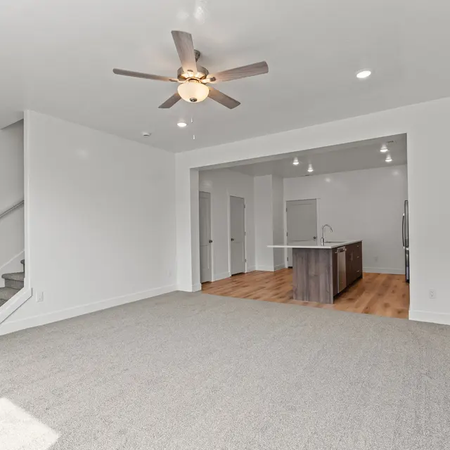 A spacious modern living room with a ceiling fan, light-colored carpet, and wooden flooring leading into an open kitchen area.