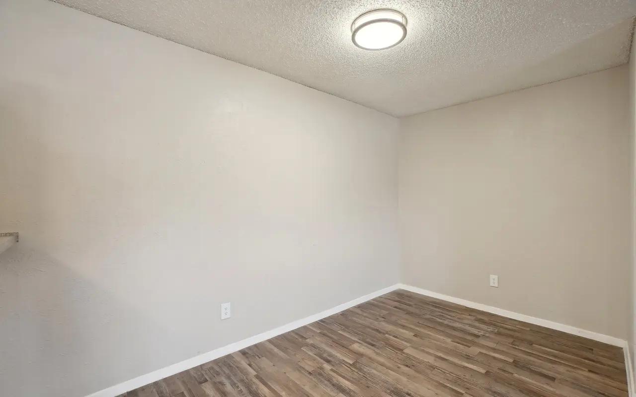 An empty room with neutral walls and wooden flooring. A ceiling light is on, illuminating the space.