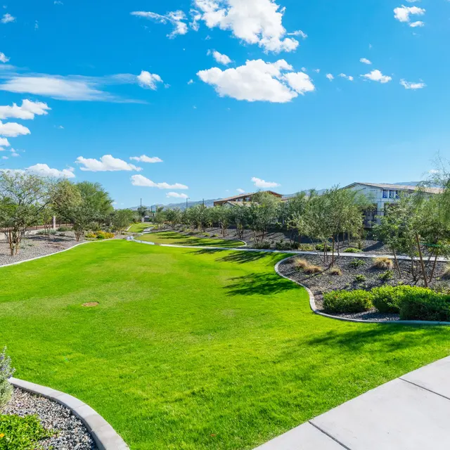 A sunny park landscape featuring a manicured green lawn bordered by gravel and dotted with trees under a blue sky with clouds.