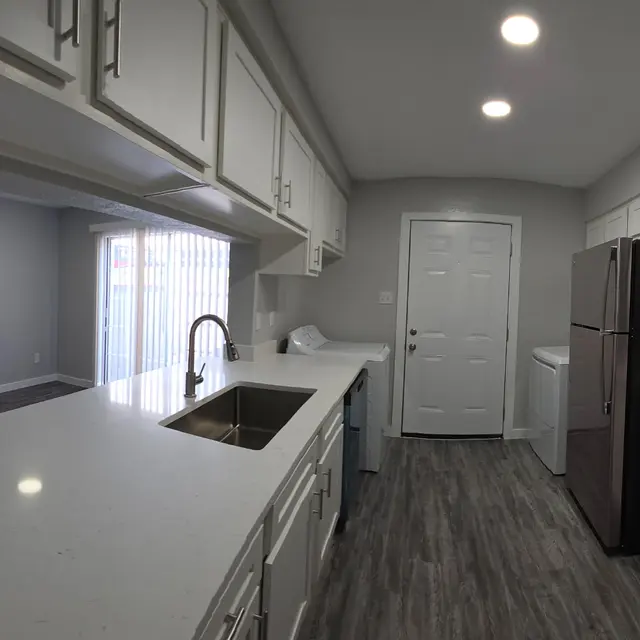 A modern kitchen featuring white cabinets, a large countertop with a sink, stainless steel appliances, and a light gray wall. The kitchen is well-lit with recessed lighting and has a spacious layout.