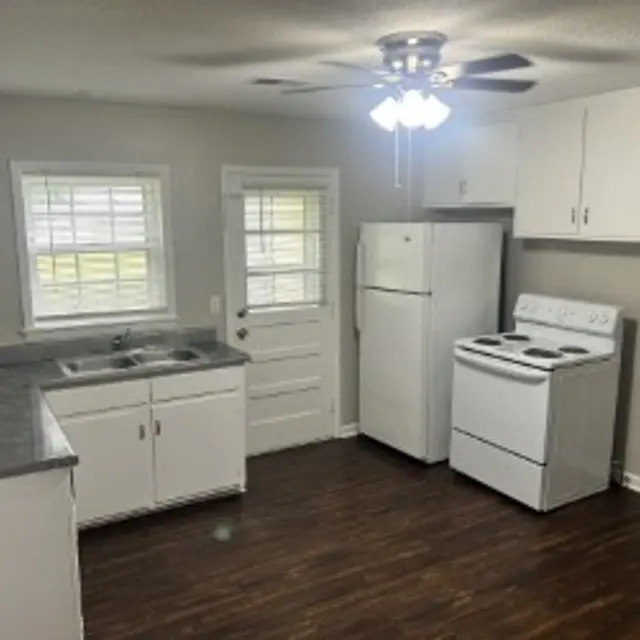A bright kitchen featuring white cabinets, a stainless-steel sink, a refrigerator, and a white stove. The kitchen has a ceiling fan and wooden flooring.