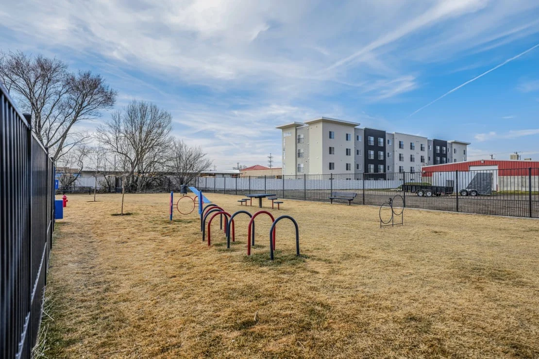 Playground Area with Buildings A playground area featuring swings and a slide, with a grassy field and a fence. In the background, several buildings including a multi-story apartment complex and a red barn-like structure are visible under a blue sky with scattered clouds.