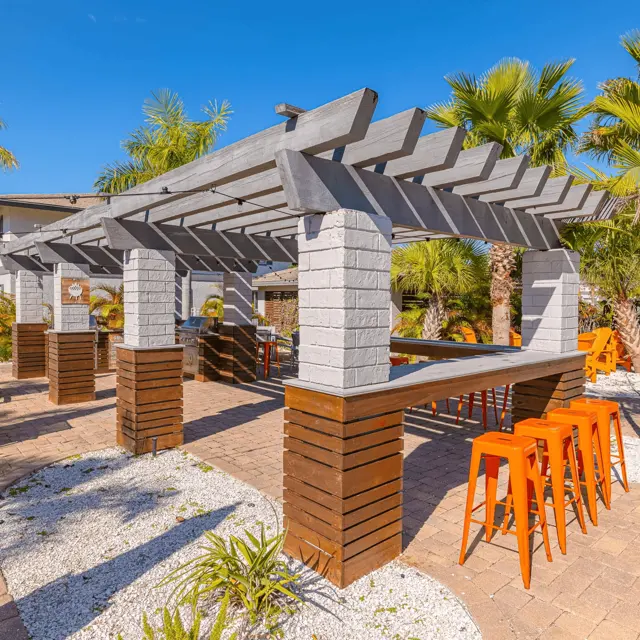 A spacious outdoor patio area featuring a pergola with tables and bright orange bar stools. Lush green palm trees and vibrant orange umbrellas add color to the setting, with a paved stone floor and white rocks providing decoration.
