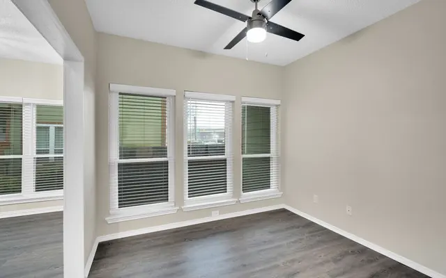 A well-lit empty room with a ceiling fan, featuring large windows with white blinds on one wall and light-colored walls. The flooring is a dark wood laminate.