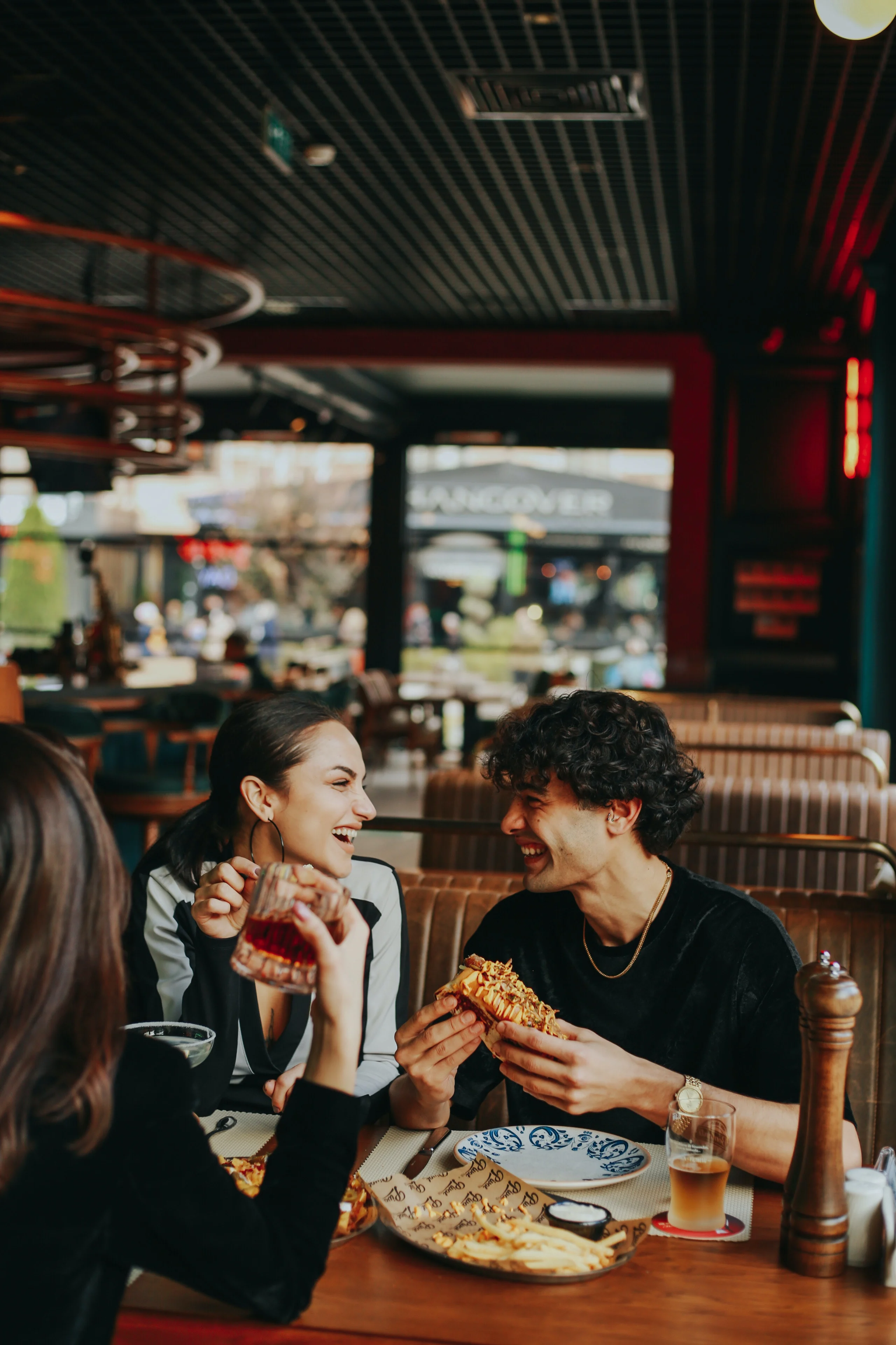 A group of friends laughing and enjoying food in a restaurant, sitting at a wooden table with a plate of pizza and drinks.