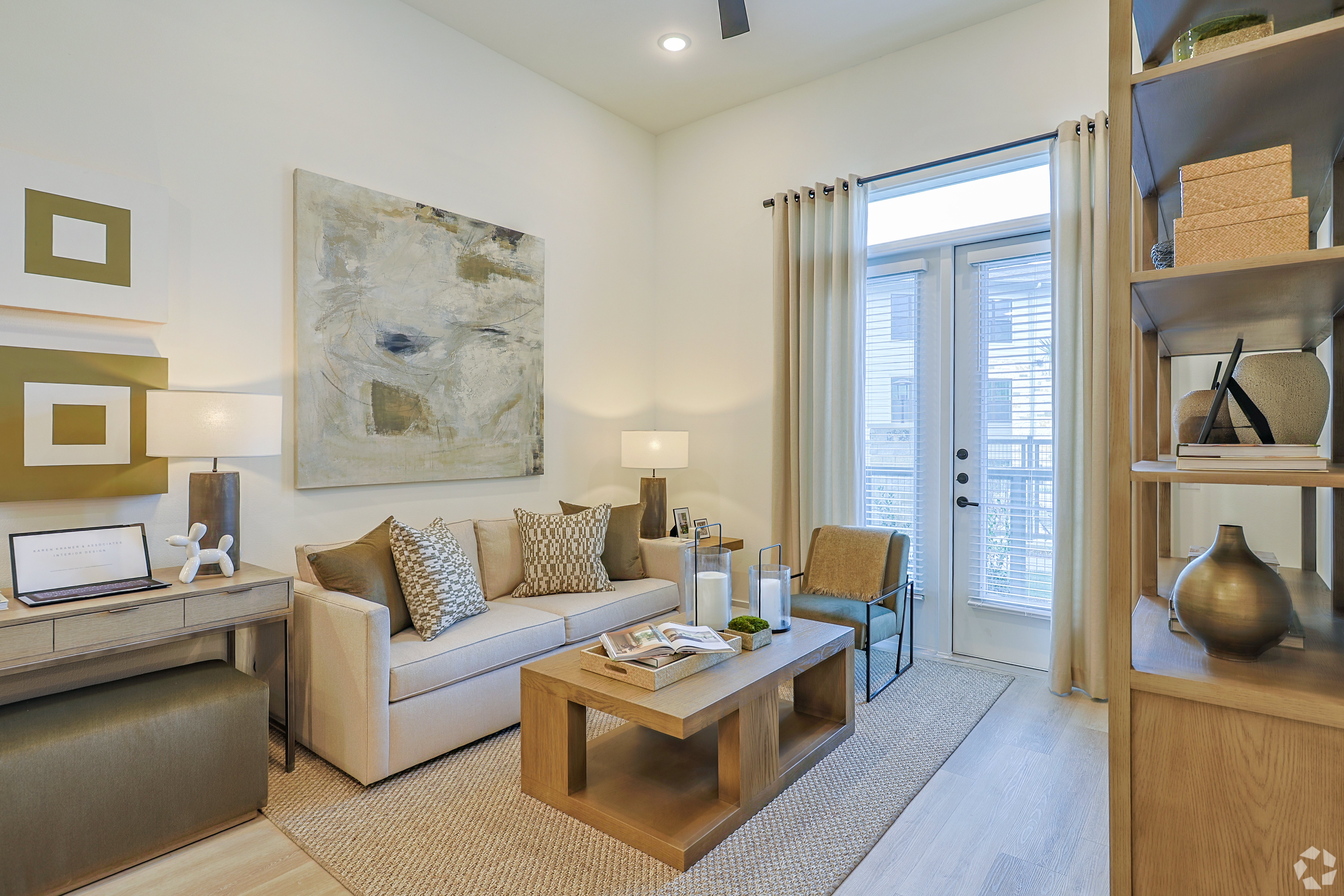 Modern Living Room Interior A modern living room featuring a light-colored sofa with decorative pillows, a wooden coffee table, and a minimalist desk. Nearby, there are large windows with sheer curtains letting in natural light, and abstract artwork on the wall. The decor includes earthy tones and light wood accents, creating a cozy atmosphere.