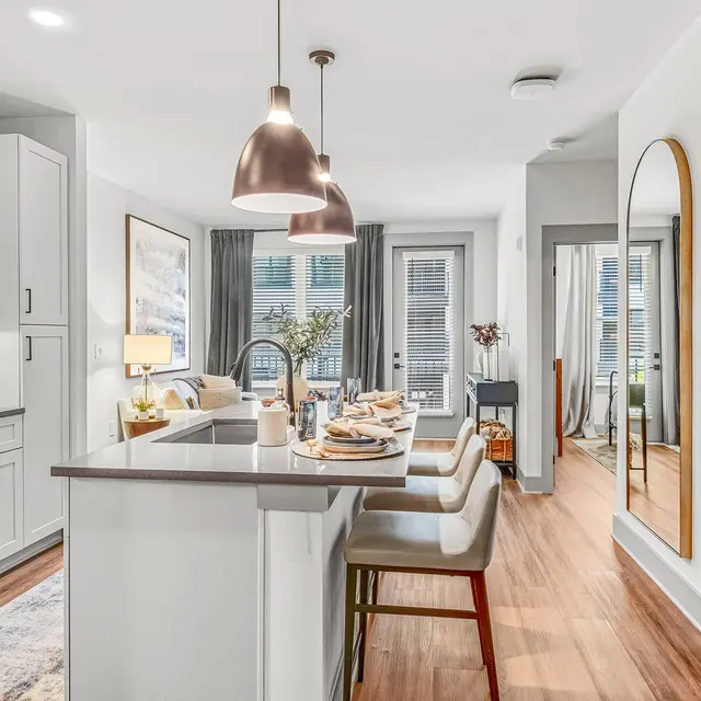 Modern Kitchen and Living Room Interior A modern kitchen and living room area featuring white cabinetry, stainless steel appliances, and pendant lighting above a breakfast bar with seating. The space has a cozy feel with decor and large windows allowing natural light in.