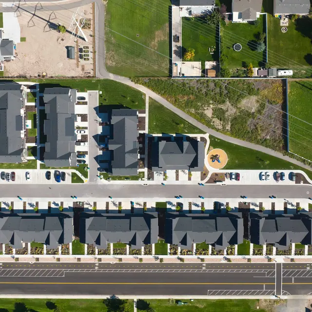Aerial view of a modern residential neighborhood featuring several rows of attached homes, green lawn areas, and parked cars. The design includes a winding road with a circular plaza.