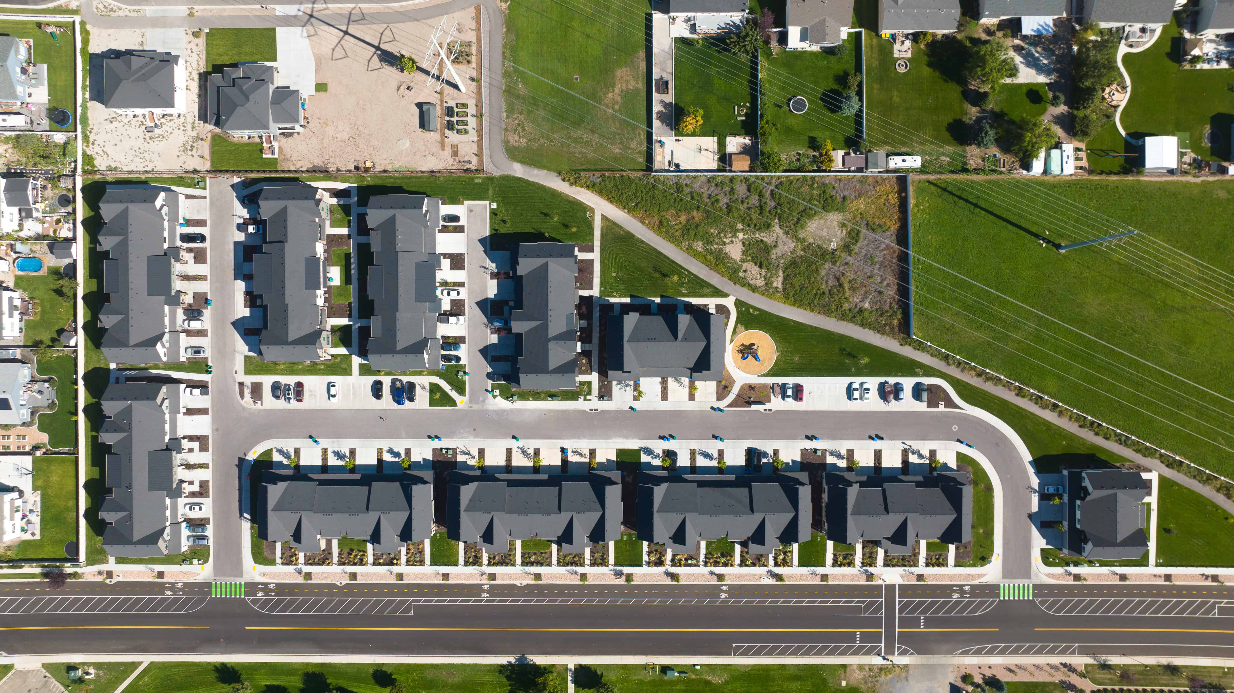 Aerial View of Residential Neighborhood Aerial view of a modern residential neighborhood featuring several rows of attached homes, green lawn areas, and parked cars. The design includes a winding road with a circular plaza.