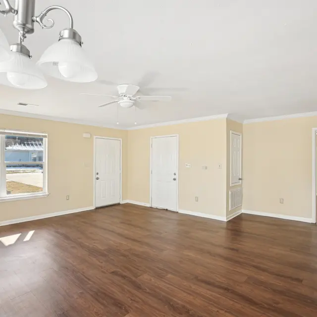 Spacious Living Room A spacious living room featuring light brown wooden flooring, a ceiling fan, and multiple windows providing natural light. There are two doors leading to different areas, and the walls are painted a soft yellow.
