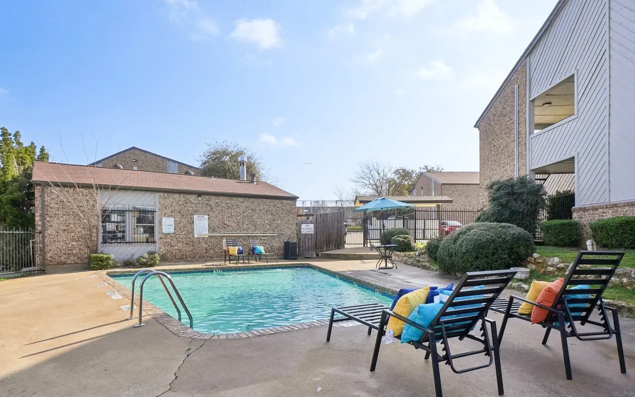 A serene pool area featuring a swimming pool surrounded by lounge chairs, colorful cushions, and greenery, with a building in the background.