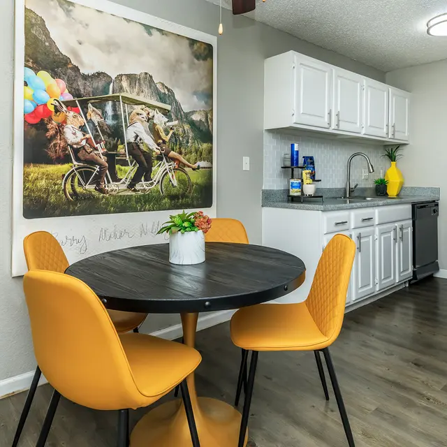 A cozy kitchen and dining area with a circular black table and four stylish yellow chairs. A vibrant wall art piece featuring a fun outdoor scene with balloons decorates the space. The kitchen is modern with white cabinets and a gray countertop.