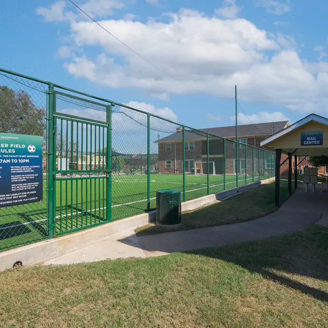 View of a soccer field enclosed by a green fence, with a pathway leading to a building. The field is well-maintained and has goal posts. There are signs indicating hours of operation.