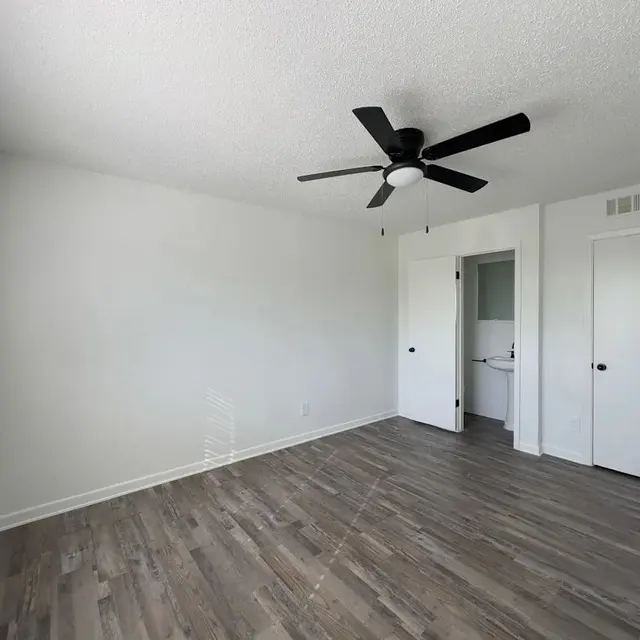 Empty Room with Ceiling Fan A bright, empty room with light-colored walls, a ceiling fan, and hardwood flooring. There are two doors leading to another room and a bathroom. A window with blinds allows natural light to enter.