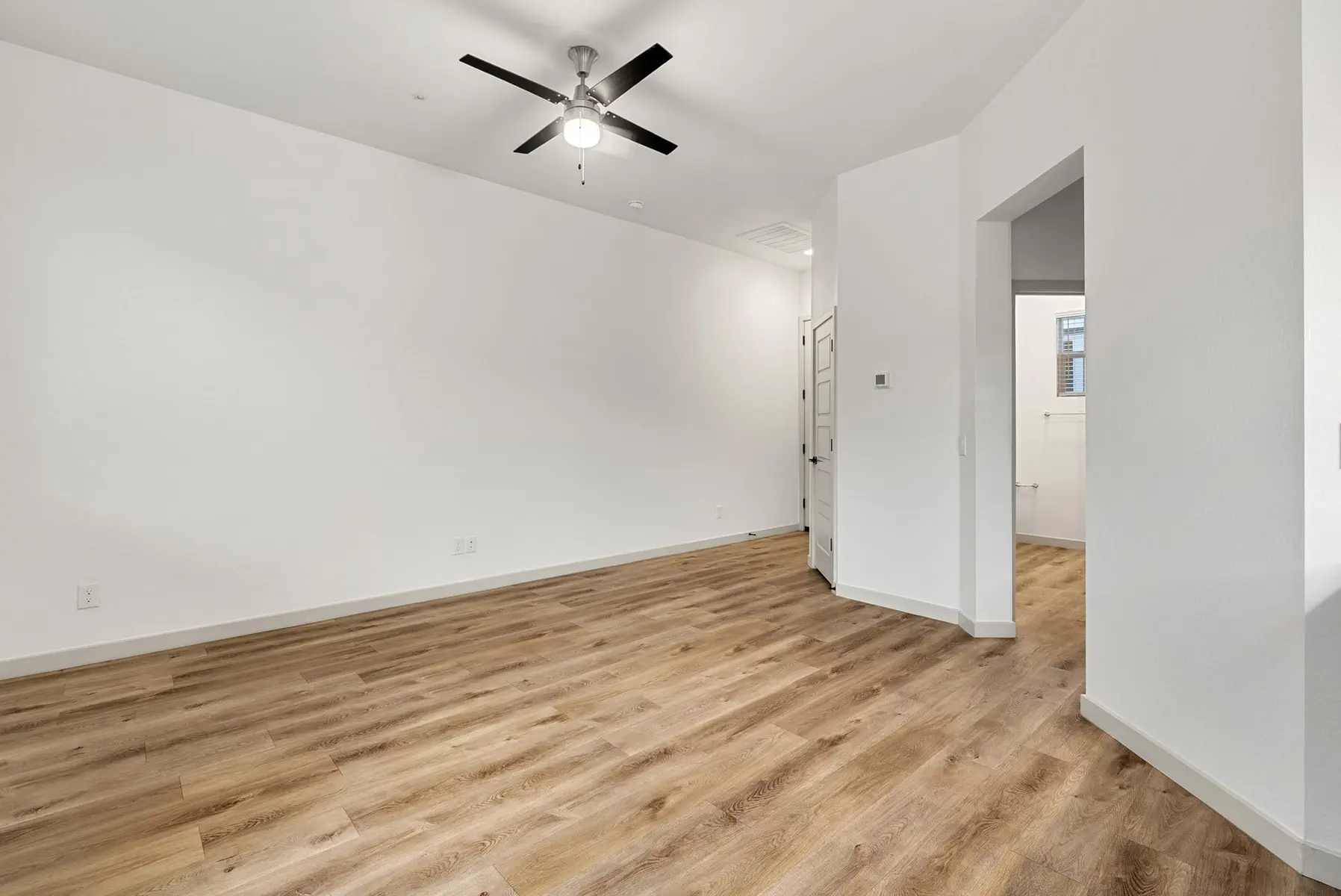 A bright, empty room with wood flooring, a ceiling fan, and white walls. An open doorway leads to another space.