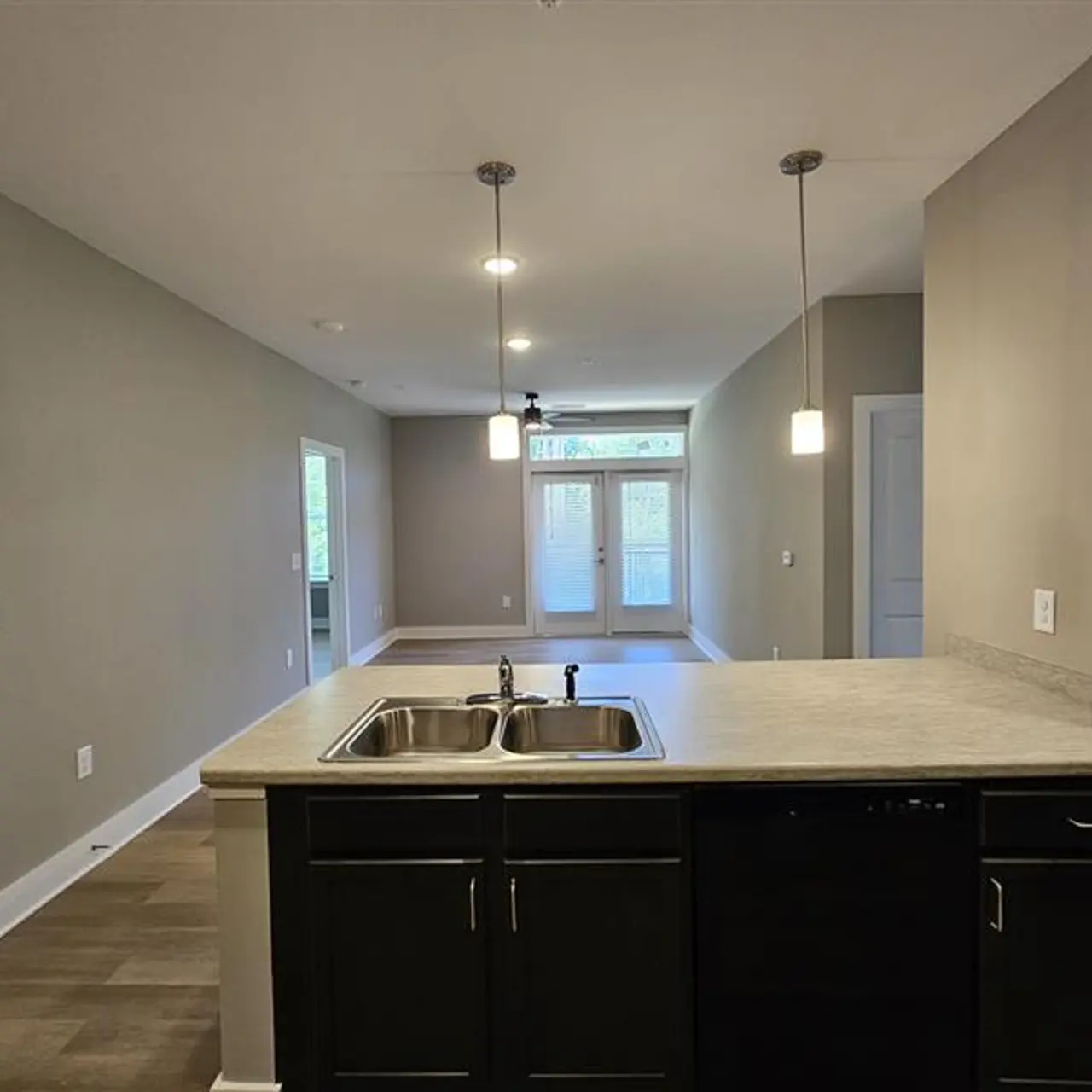 A view of a modern kitchen with a dark countertop, double sink, and black appliances. The kitchen opens to a spacious living area with neutral walls and natural light coming through double doors at the back.