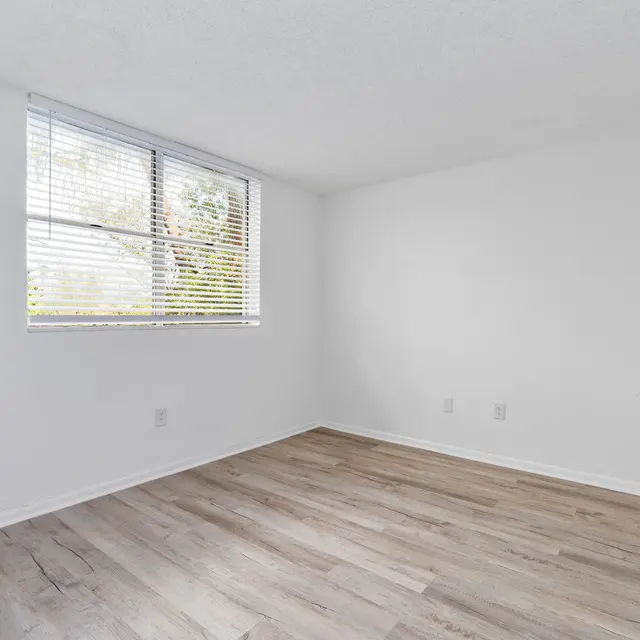 A vacant room with light-colored walls and wooden flooring. There is a window with blinds allowing natural light to enter. The room appears clean and freshly painted with no furniture.