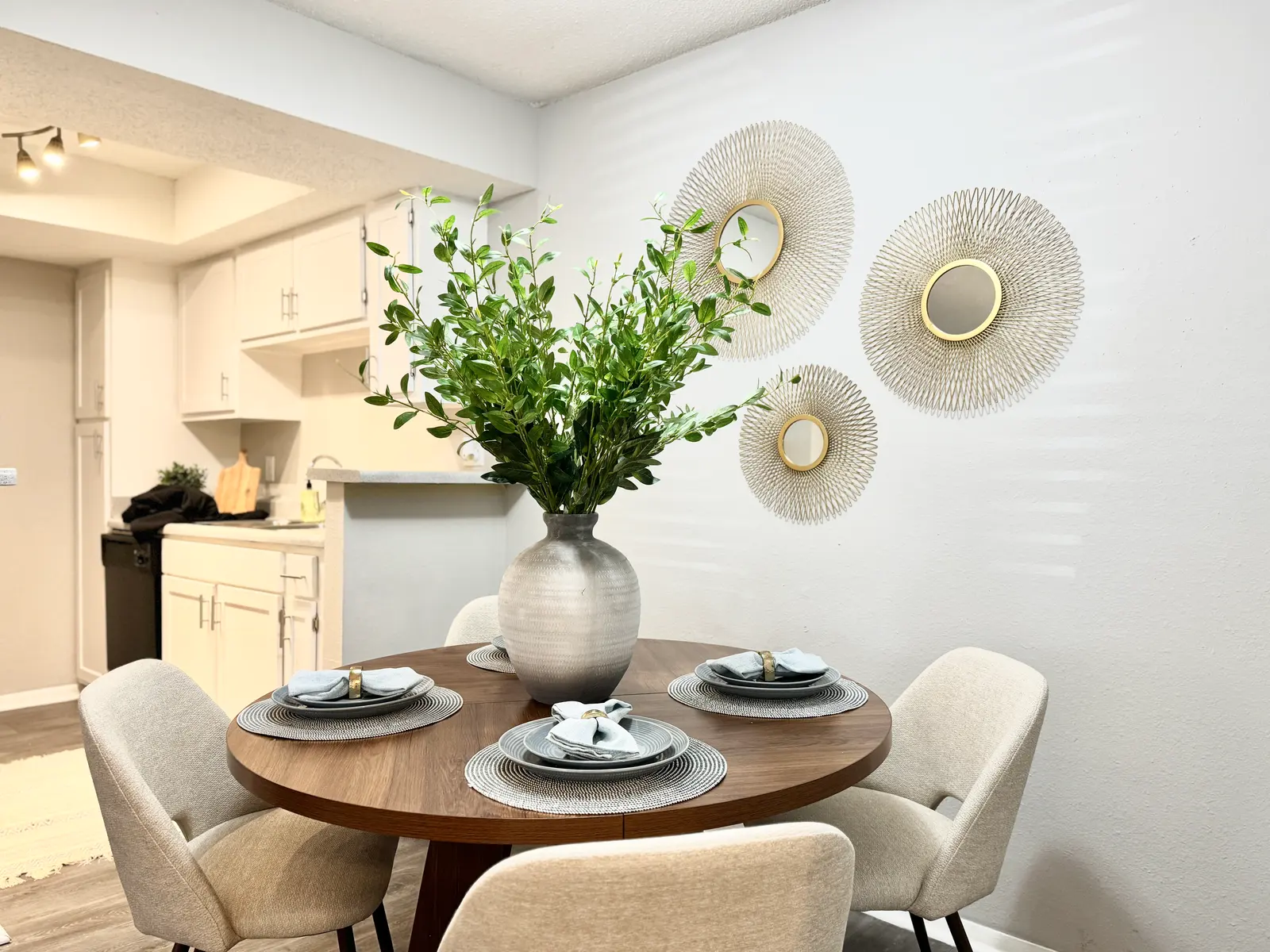 A modern dining area featuring a round wooden table with four upholstered chairs. A large vase with greenery is in the center of the table, accompanied by stylish table settings. Three decorative wall plates are mounted on the light gray wall behind the table.