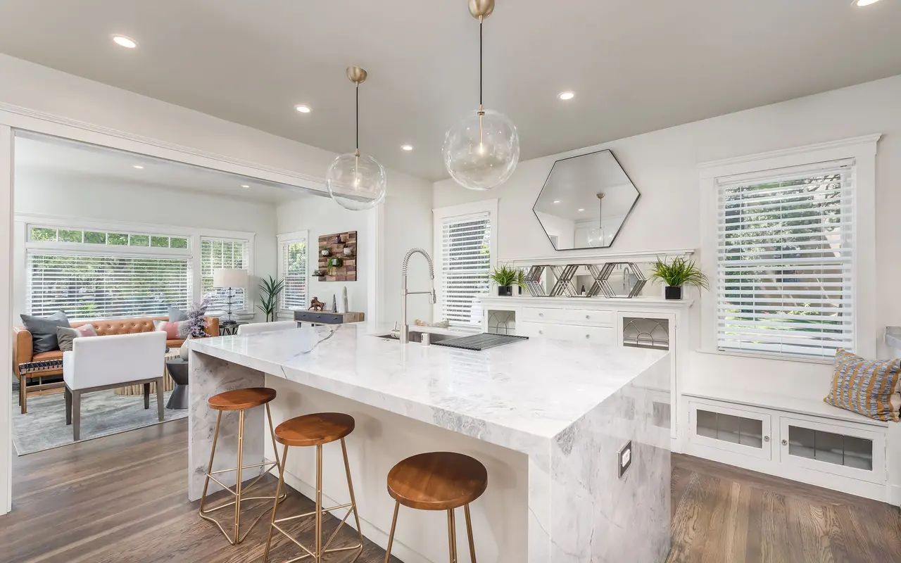 A modern kitchen featuring a large marble island with three wooden bar stools, pendant lighting, and a stylish faucet. In the background, a cozy living area with windows and plants can be seen.